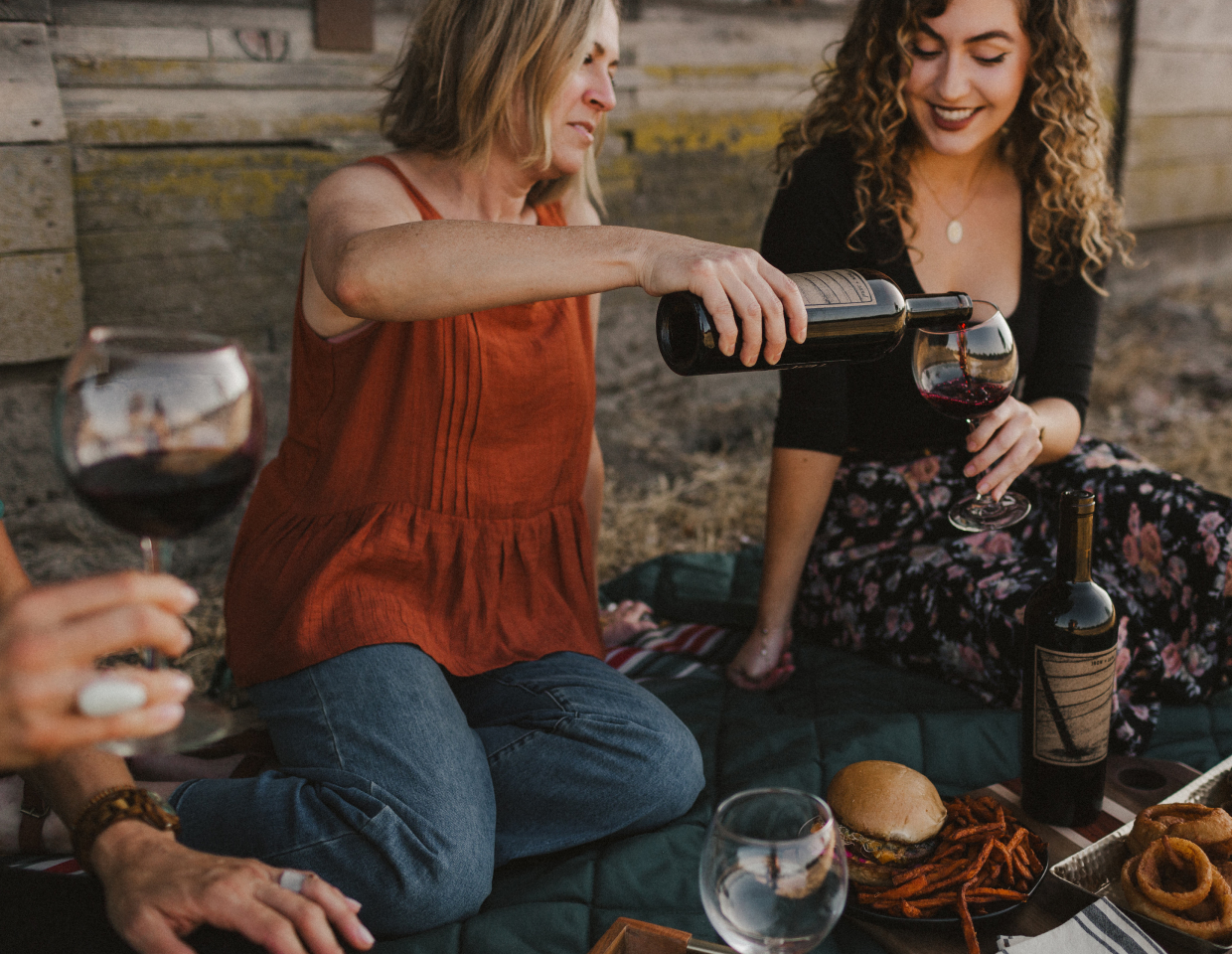 Women sitting on a picnic blanket with burgers and fries, pouring a glass of Iron + Sand cabernet sauvignon.
