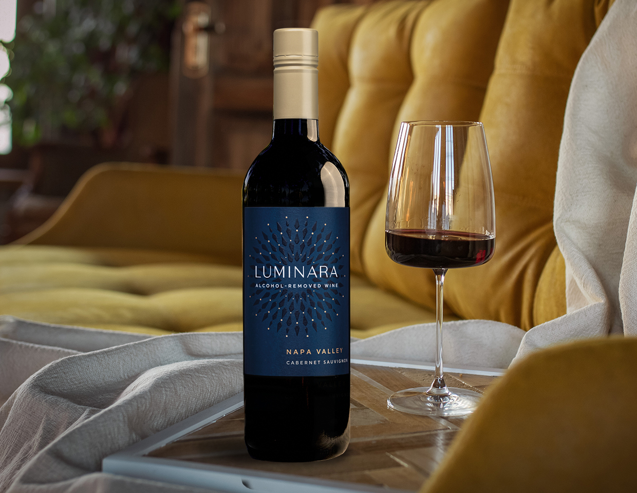 bottle and glass of luminara alcohol-removed caberet sauvignon on a table beside a yellow couch