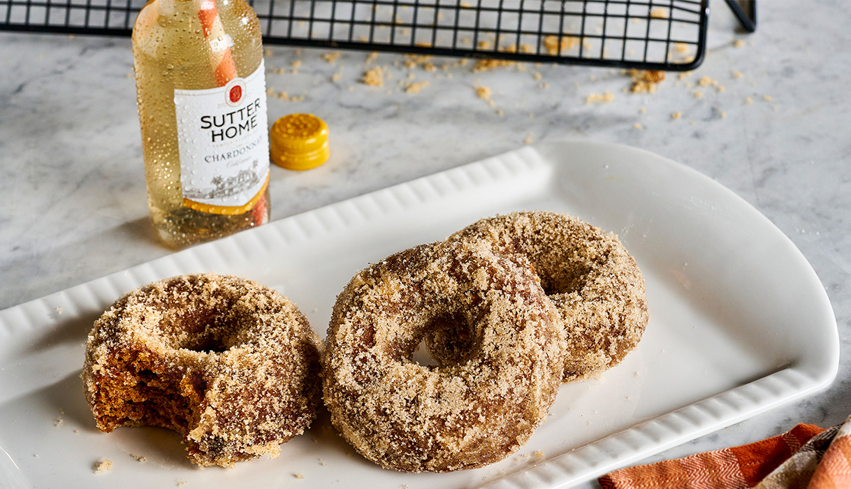 Three apple cider donuts, one with a bite taken out of it, on a white serving platter with a small bottle of Sutter Home Chardonnay in the background