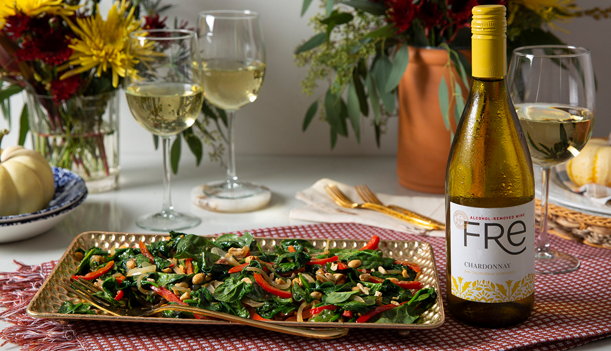 Stir friend spinach and peanuts on a platter next to a bottle and glasses of FRE alcohol-removed chardonnay on a gingham placemat with fall flowers in the background.
