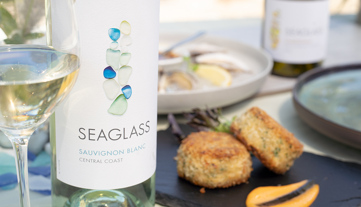 crab cakes on a plate next to a bottle and glass of SEAGLASS central coast sauvignon blanc with more seafood and wine in the background.
