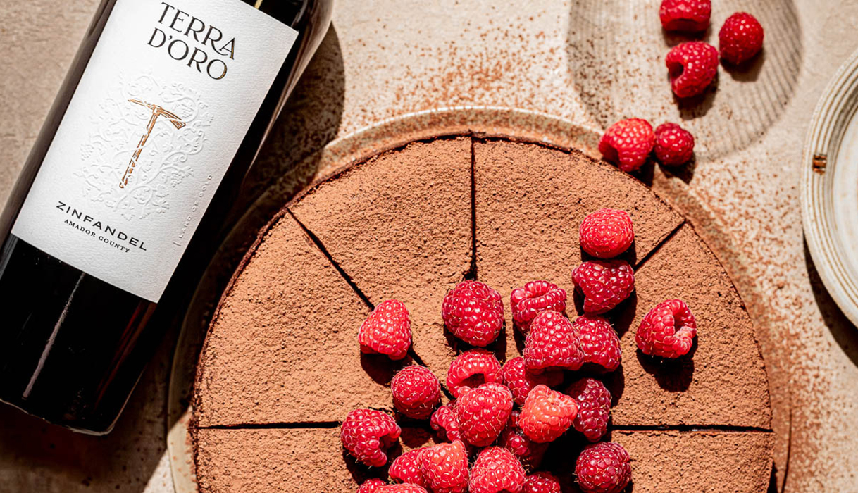 Bottle of Terra d'Oro zinfandel lying next to a red wine chocolate cake with a pile of fresh raspberries on top trailing onto the counter.