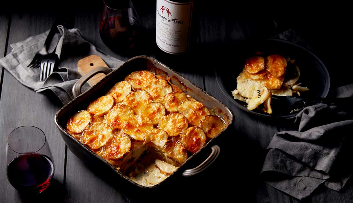 a dish of Tartiflette or cheesy potato casserole with a slice cut out and placed on a black plate, a bottle and glasses of menage a trois red blend are in the background.